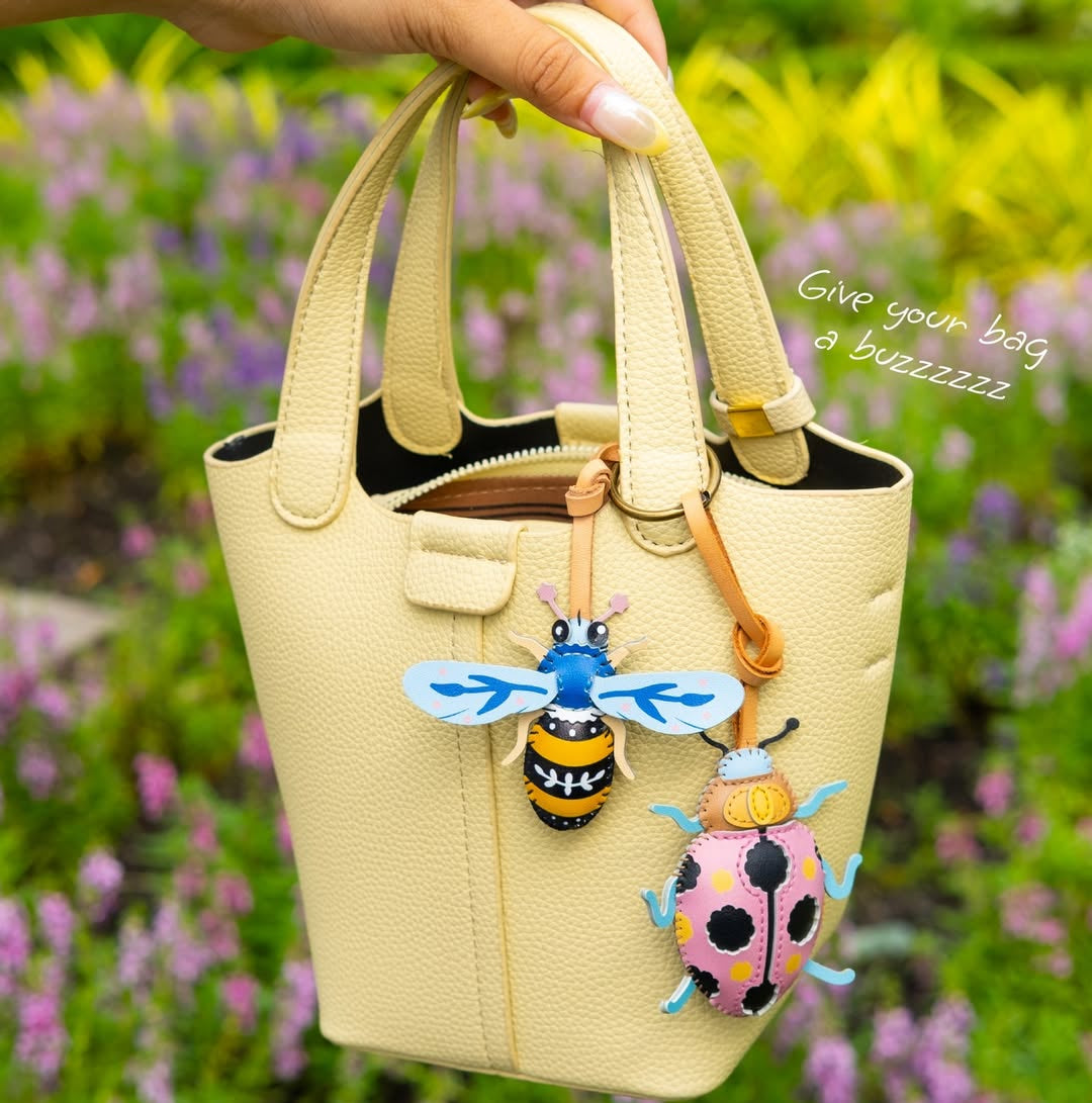 Beige handbag with bee and ladybug keychains held by a hand against a floral background.